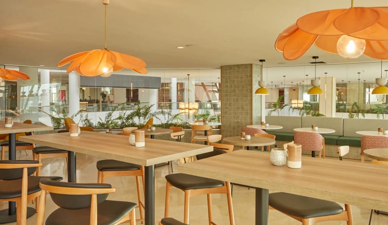 Coco Cafe Bright and modern resort café seating area featuring light wood tables, black cushioned chairs, and large orange petal-shaped pendant lights. at Dreams Lanzarote Playa Dorada Resort & Spa by Kenwood Travel