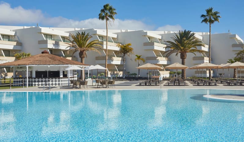 Barracuda pool bar with a thatched roof, white multi-story hotel buildings, and palm trees under a blue sky at Dreams Lanzarote Playa Dorada Resort & Spa by Kenwood Travel