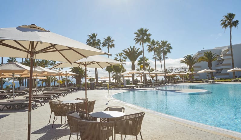 Barracuda featuring woven outdoor dining chairs, white patio umbrellas, and sun loungers surrounding a large blue swimming pool with palm trees at Dreams Lanzarote Playa Dorada Resort & Spa by Kenwood Travel