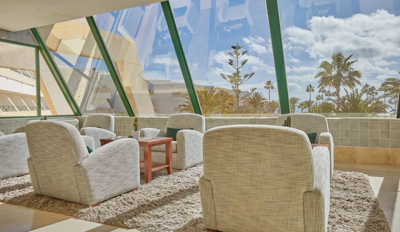 Preferred Club lounge with comfortable grey armchairs on a plush rug, featuring large slanted windows with views of palm trees and a bright blue sky at Dreams Lanzarote Playa Dorada Resort & Spa by Kenwood Travel