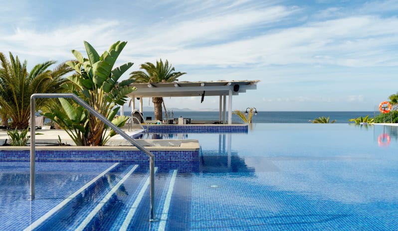 Sea Legs Infinity pool with blue mosaic tiles overlooking the ocean, featuring a poolside bar and tropical palm trees under a bright sky at Dreams Lanzarote Playa Dorada Resort & Spa by Kenwood Travel