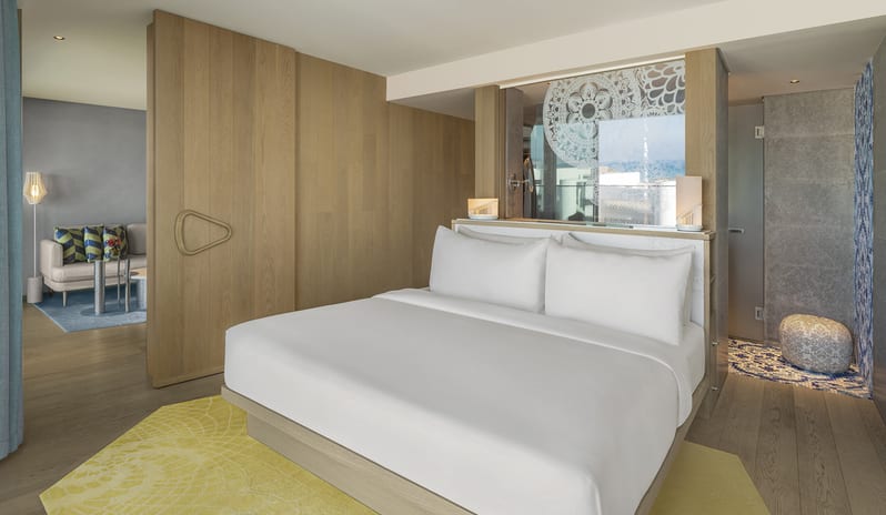 Premier Two Bedroom Suite king-sized bed with white linens, a yellow patterned rug, light wood paneling, and a glimpse into a connected living area with a sofa at W Algarve by Kenwood Travel