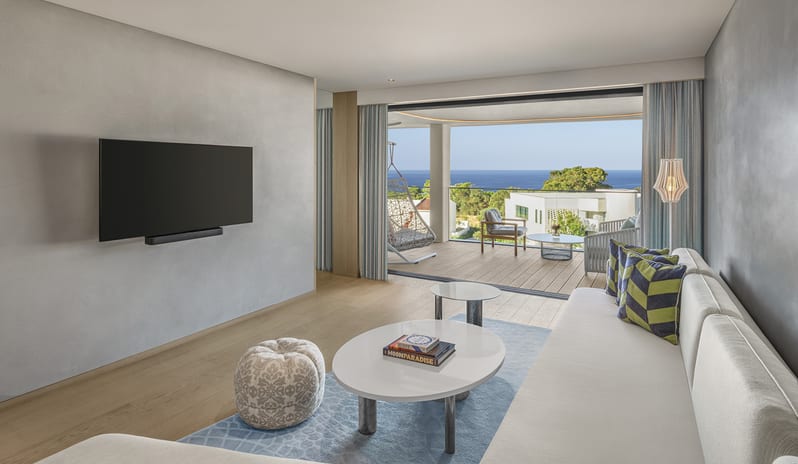 Premier Two Bedroom Suite featuring a light gray sofa, wall-mounted TV, and large sliding doors opening onto a private wooden balcony with an ocean view at W Algarve by Kenwood Travel