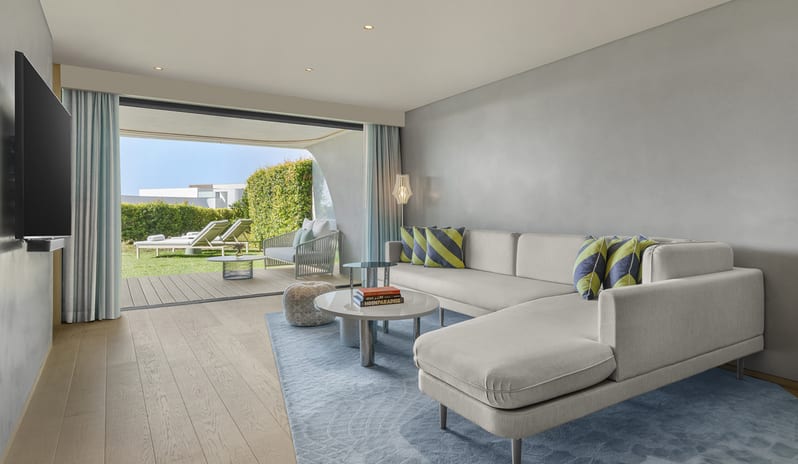 Superior Two Bedroom Suite featuring a light gray L-shaped sofa, blue rug, and floor-to-ceiling glass doors opening to a private terrace with lounge chairs at W Algarve by Kenwood Travel