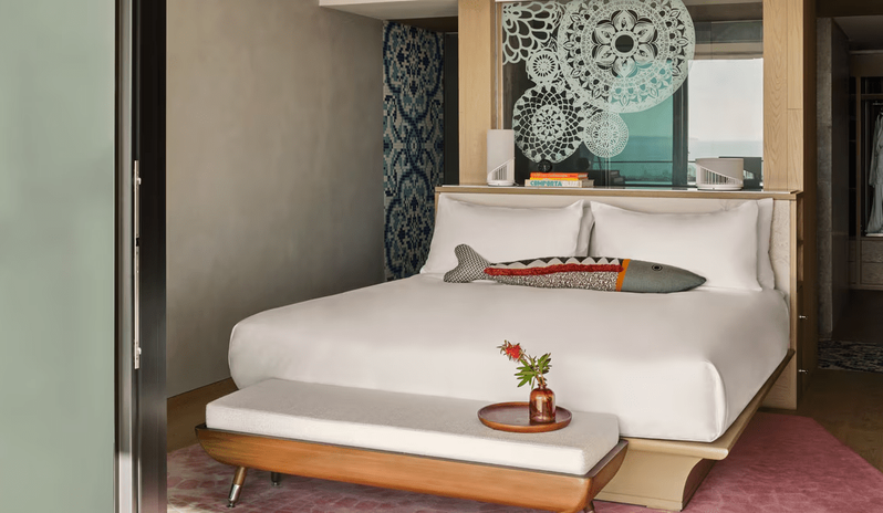 Supreme Two-Bedroom Residence bedroom featuring a white platform bed with a decorative fish pillow, a wooden end-of-bed bench, and a glass partition with white mandala patterns overlooking the ocean at W Algarve by Kenwood Travel