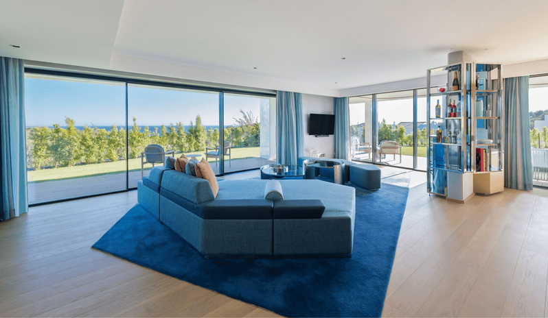 Supreme Two-Bedroom Residence modern blue modular sofa, a breakfast bar with stools, and large floor-to-ceiling sliding glass doors opening onto a private green lawn with a hanging swing chair at W Algarve by Kenwood Travel