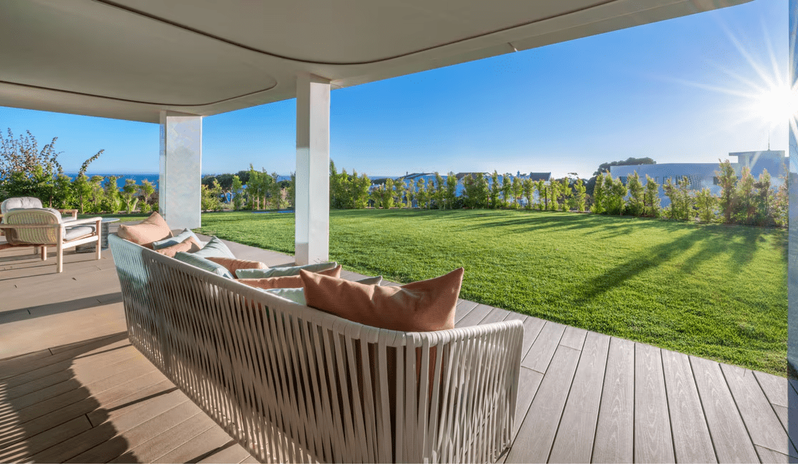 Supreme Two-Bedroom Residence outdoor terrace with a stylish woven sofa and colorful pillows, overlooking a lush green lawn and garden under a bright sunny sky with a sunburst effect at W Algarve by Kenwood Travel