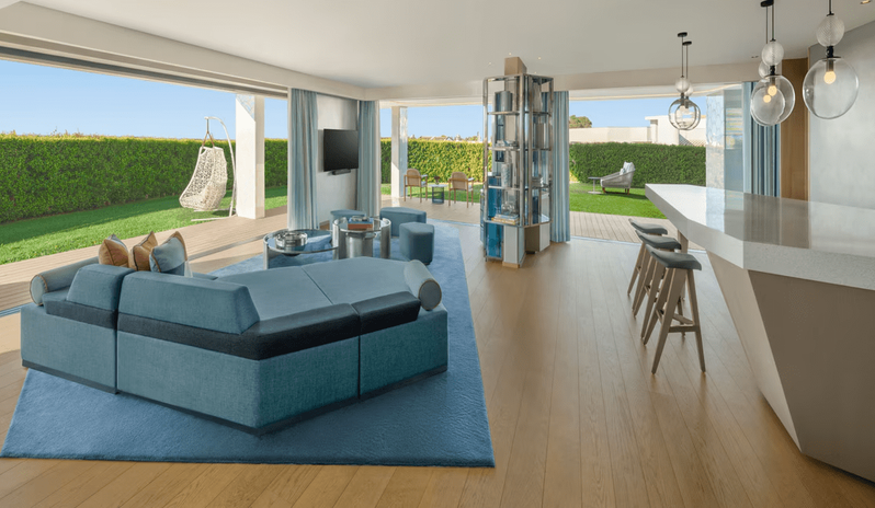 Supreme Two-Bedroom Residence modern blue modular sofa, a breakfast bar with stools, and large floor-to-ceiling sliding glass doors opening onto a private green lawn with a hanging swing chair at W Algarve by Kenwood Travel