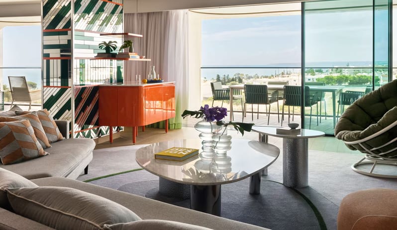 Supreme Three-Bedroom Residence Sun-lit modern living room with a bright orange credenza, a decorative geometric glass partition, grey sectional sofa, and marble coffee tables overlooking a scenic balcony at W Algarve by Kenwood Travel
