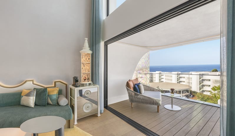 W Suite with Plunge Pool, One Bedroom Suite living area of a luxury hotel suite featuring a green velvet sofa, a mirrored bar cart, and large sliding glass doors opening onto a private wooden balcony with a wide ocean view at W Algarve by Kenwood Travel