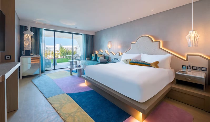 Premier Private Garden Room featuring a large bed with a decorative fish pillow, a vibrant multicolored area rug, and sliding glass doors opening onto a sunny terrace at W Algarve by Kenwood Travel