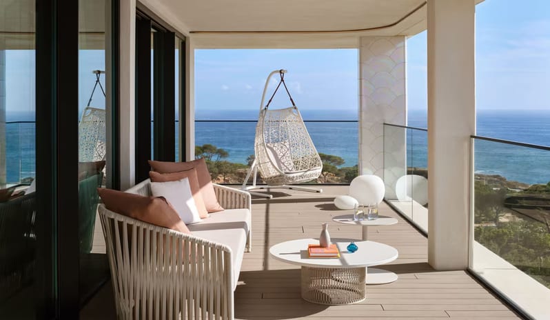 Penthouse I, Two Bedroom Suite with Pool balcony with a panoramic ocean view, featuring a white woven outdoor sofa with salmon pillows, a hanging egg chair, and modern round coffee tables at W Algarve by Kenwood Travel