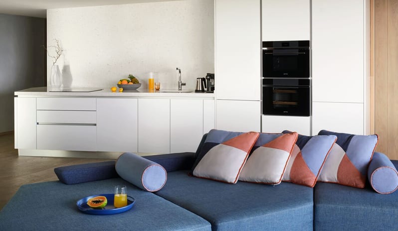 Penthouse I, Two Bedroom Suite with Pool kitchenette with white handleless cabinetry and built-in black ovens, positioned behind a blue modular sofa featuring geometric-patterned cushions at W Algarve by Kenwood Travel
