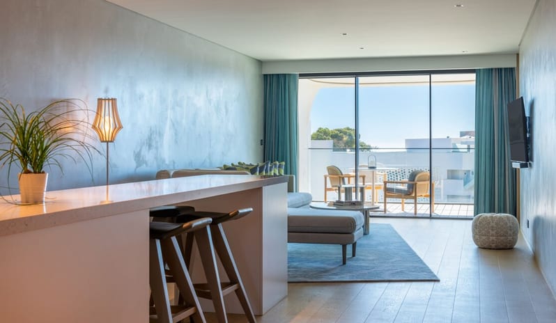Premier Two-Bedroom Residence living room at W Algarve, featuring a sleek white breakfast bar with stools, a modern grey sofa, and floor-to-ceiling glass doors opening onto a sunlit private balcony with outdoor seating at W Algarve by Kenwood Travel