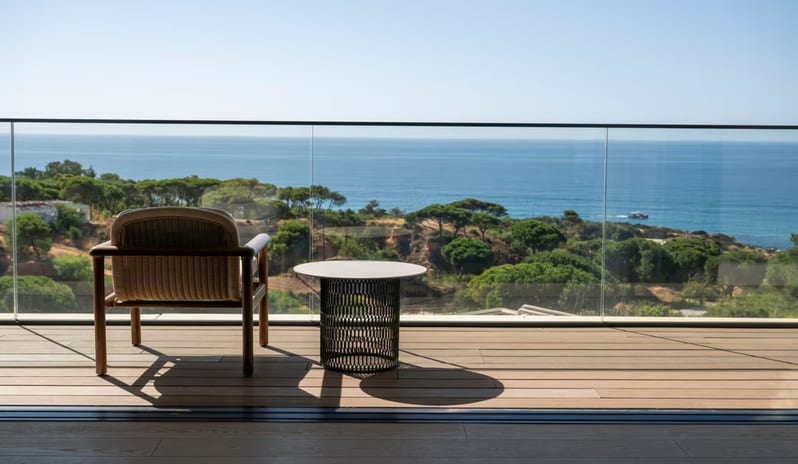 Penthouse I, Two Bedroom Suite with Pool rooftop tbalcony with wood-plank flooring and a glass railing, featuring a contemporary woven armchair and a small round black side table overlooking the ocean and coastal trees at W Algarve by Kenwood Travel