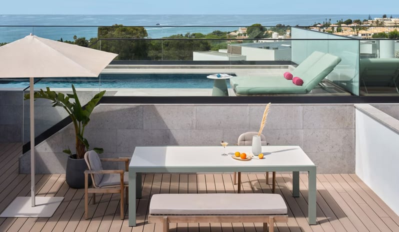 Supreme Two Bedroom Penthouse with Pool rrooftop terrace featuring a minimalist outdoor dining set with a bench, a tan patio umbrella, and a glass-walled infinity pool overlooking the ocean at W Algarve by Kenwood Travel