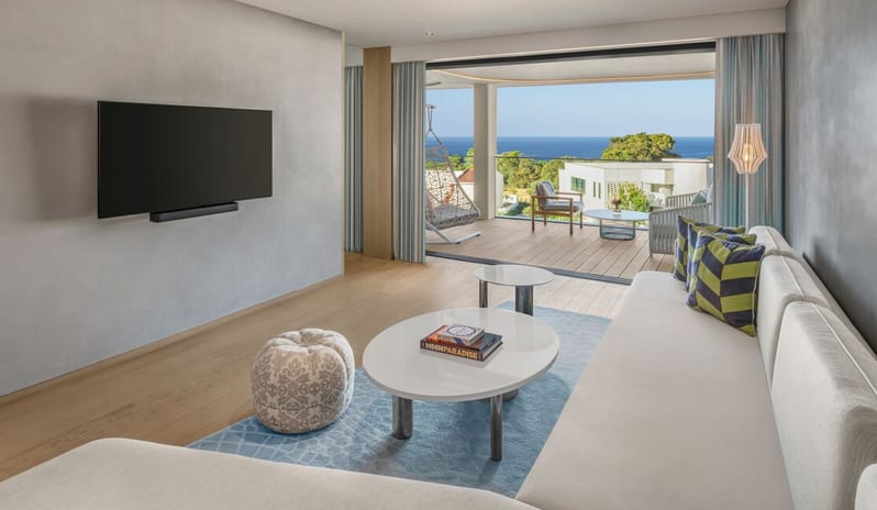 Premier Two-Bedroom Residence lliving room in a Premier Suite at W Algarve, featuring a neutral sectional sofa, a wall-mounted TV, and wide sliding doors that open onto a wooden deck terrace with a hanging chair and panoramic ocean views at W Algarve by Kenwood Travel