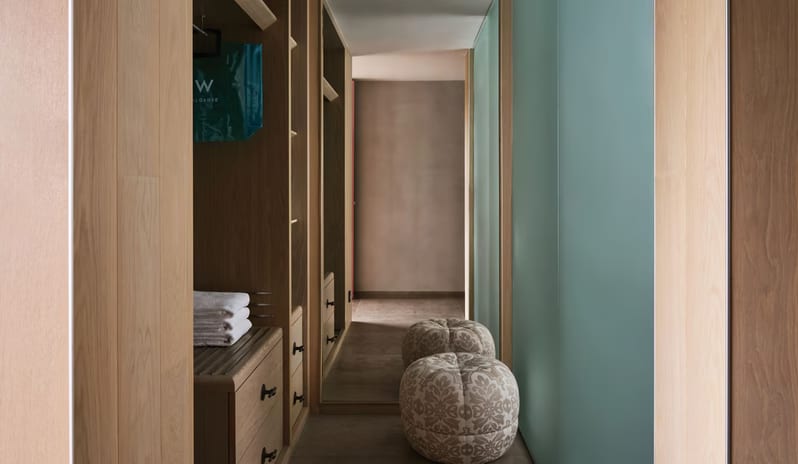 Premier Private Garden Room modern walk-in closet in a luxury hotel room featuring light wood cabinetry, a stack of white towels, and two decorative patterned floor poufs in a narrow hallway at W Algarve by Kenwood Travel