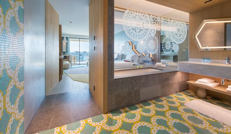Premier Deluxe Room bathroom with vibrant teal and yellow mosaic floor tiles, a deep soaking tub next to a glass partition with white mandala decals, and a sleek marble vanity with a geometric backlit mirror at W Algarve by Kenwood Travel