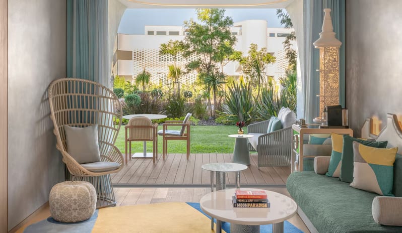 Superior Room living area with a teal sofa and high-back wicker chair, opening onto a wooden ground-floor terrace with outdoor seating and a lush green garden view. at W Algarve by Kenwood Travel