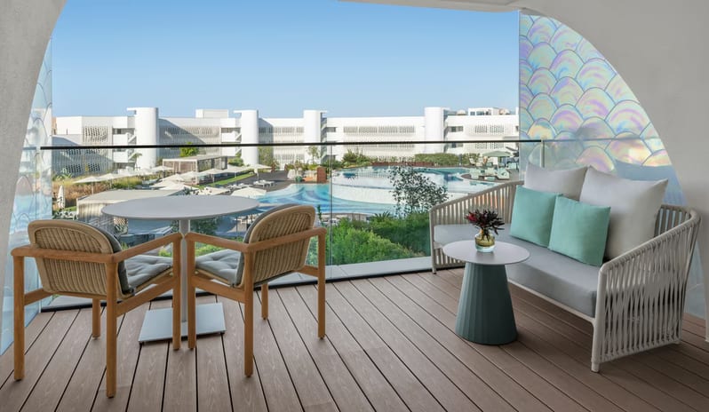 Superior Room terrace balcony with a modern dining set and a gray lounge sofa with teal cushions, featuring a glass railing that overlooks a large resort swimming pool and white architectural buildings at W Algarve by Kenwood Travel
