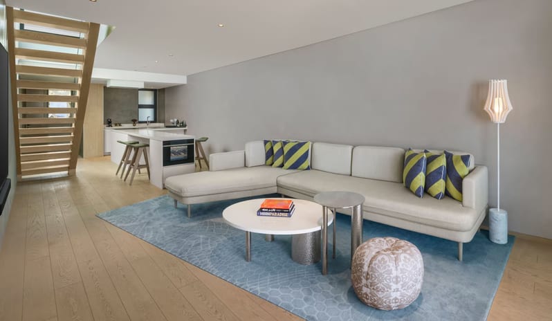 Supreme Two Bedroom Penthouse with Pool living area with an open-plan layout, featuring a large cream sectional sofa with striped pillows, a light blue textured rug, and a minimalist kitchen in the background with a wooden staircase at W Algarve by Kenwood Travel