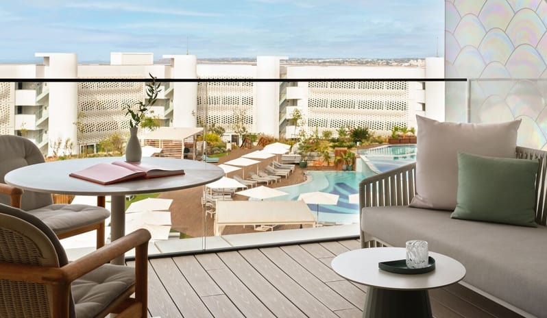 Superior Room balcony featuring a wooden deck, a white marble bistro table, and comfortable outdoor lounge seating, overlooking a landscaped swimming pool area with white umbrellas at W Algarve by Kenwood Travel