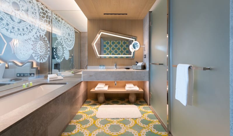 Superior Room bathroom featuring a floating marble vanity with an asymmetrical backlit mirror, vibrant yellow and teal patterned floor tiles, and a large soaking tub next to a decorative glass partition at W Algarve by Kenwood Travel