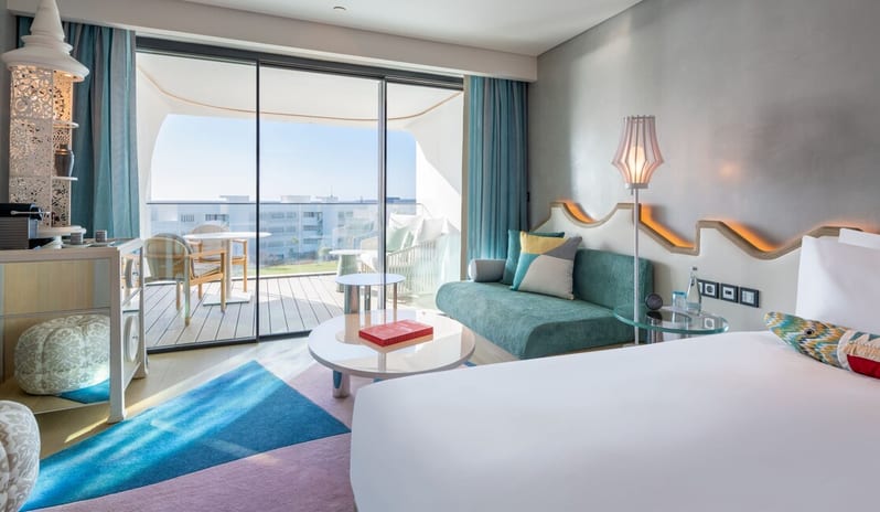 Superior Room living area featuring a teal sofa, a round white coffee table, and large sliding glass doors that open onto a private balcony with outdoor seating and a coastal view at W Algarve by Kenwood Travel