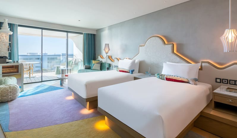 Premier Deluxe Room featuring two beds with glowing under-lighting, a decorative white headboard, colorful geometric rug, and a private balcony with a sea view at W Algarve by Kenwood Travel