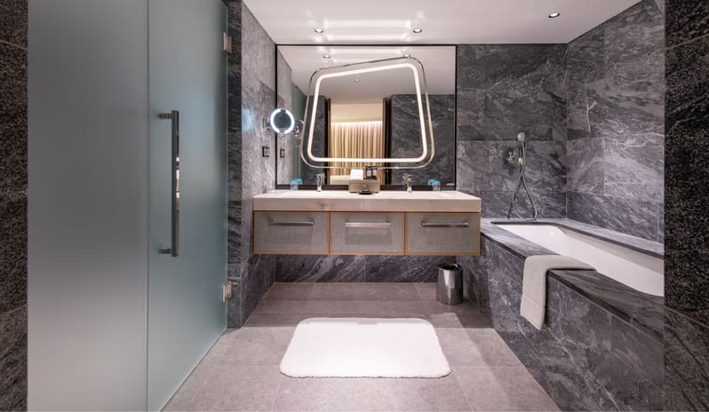 Premier One-Bedroom Residence bathroom featuring dark grey marble walls, a double vanity with a backlit geometric mirror, a white soaking tub, and a frosted glass shower door at W Algarve by Kenwood Travel