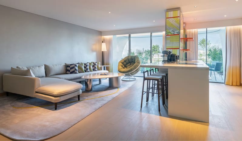 Premier One-Bedroom Residence open-plan hotel suite featuring a grey sectional sofa with patterned cushions, a white kitchen island with bar stools, and a round green swivel chair next to floor-to-ceiling window at W Algarve by Kenwood Travel