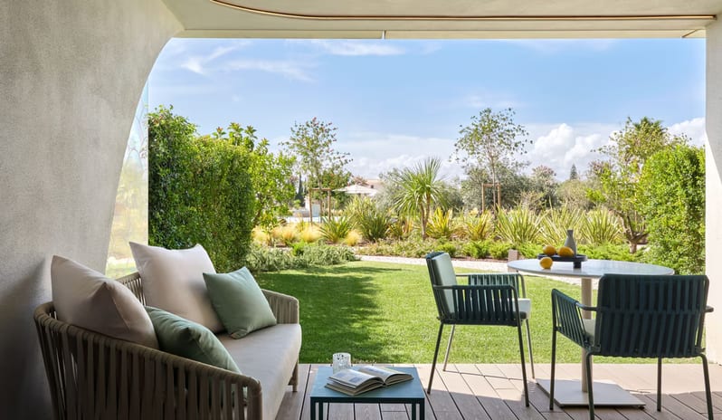 Premier One-Bedroom Residence terrace with modern outdoor lounge furniture, including a sofa with sage green pillows and a dining table, overlooking a lush landscaped garden with green lawns and palm trees under a bright blue sky at W Algarve by Kenwood Travel