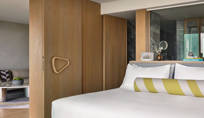 Supreme Two Bedroom Suite featuring a light wood sliding pocket door with a triangular handle, a white bed with a striped bolster pillow, and a glimpse into a living area at W Algarve by Kenwood Travel