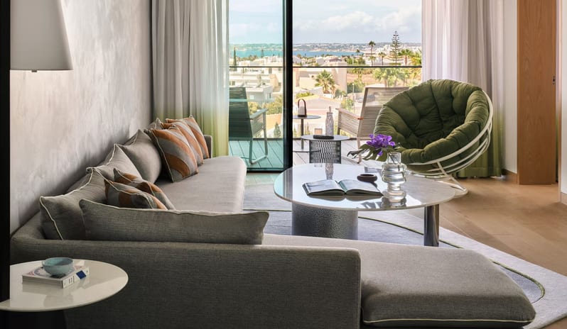 Supreme Two Bedroom Suite living room featuring a grey sectional sofa with orange patterned pillows, a marble coffee table, and a green papasan chair leading to a private balcony with a coastal city view at W Algarve by Kenwood Travel