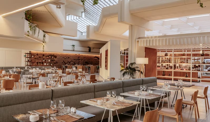 Malva Food Bazaar Restaurant interior featuring a large backlit stone relief wall mural, modern teal rope chairs, and elegantly set tables with wine glasses and gourmet plates at Paradisus Salinas Lanzarote by Kenwood Travel