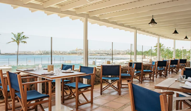 La Graciosa Restaurant beachfront dining terrace at La Graciosa in Lanzarote featuring wooden tables, navy blue chairs, and a glass railing overlooking the ocean and coast at Paradisus Salinas Lanzarote by Kenwood Travel