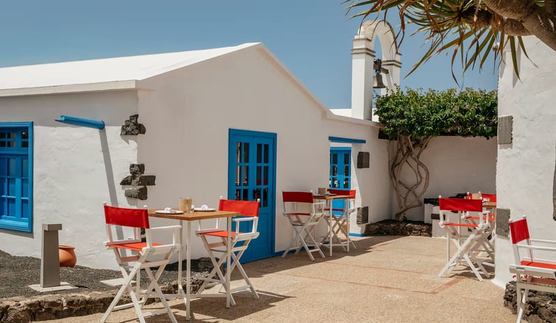 La Graciosa Restaurant Traditional white-washed outdoor patio at La Graciosa in Lanzarote featuring blue window frames, red director chairs, and a small bell tower under a bright sunny sky at Paradisus Salinas Lanzarote by Kenwood Travel