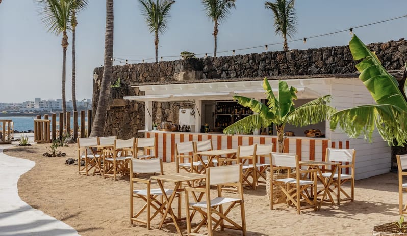 Beach Bar with white and orange striped counter, wooden director chairs, and palm trees against a volcanic stone wall at Paradisus Salinas Lanzarote by Kenwood Travel
