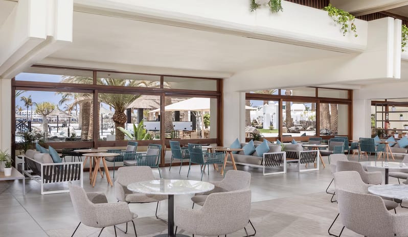 Ginger Lobby Bar lounge with marble tables, teal wicker chairs, and large glass windows overlooking a tropical resort with palm trees at Paradisus Salinas Lanzarote by Kenwood Travel