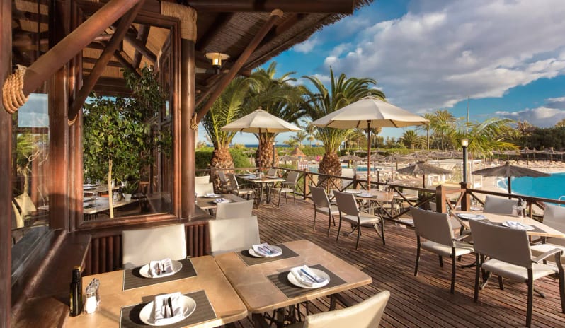 La Veranda Restaurant restaurant featuring set tables, white umbrellas, and palm trees overlooking a resort swimming pool under a blue sky at Sheraton Fuerteventura Beach, Golf & Spa Resort by Kenwood Travel