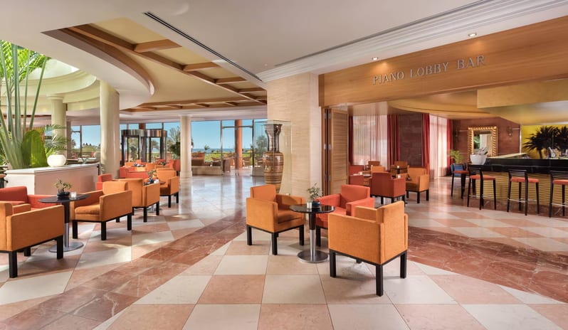 Piano Lobby Bar Spacious luxury hotel lobby bar with orange armchairs, marble checkered flooring, and a view of a tropical garden at Sheraton Fuerteventura Beach, Golf & Spa Resort by Kenwood Travel