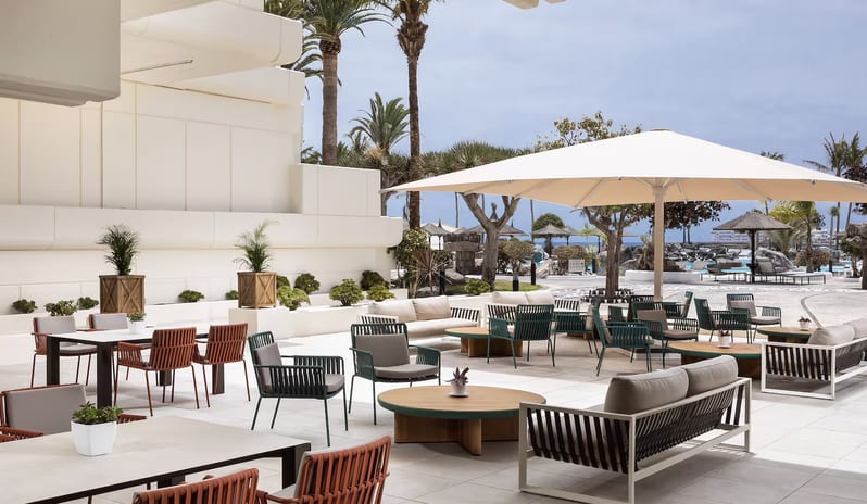 The Shack Poolside Terrace  resort featuring round wooden coffee tables, and a large white cantilever umbrella overlooking a poolside area with palm trees at Paradisus Salinas Lanzarote by Kenwood Travel