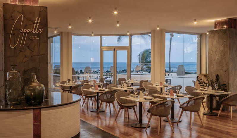 Cappella Italian Restaurant dining room with marble tables, woven chairs, and a view of the sea through large glass doors at Paradisus Salinas Lanzarote by Kenwood Travel