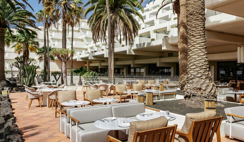 Samira restaurant featuring white stepped architectural balconies, tall palm trees, and modern grey lounge seating with marble tables set against a volcanic rock landscape at Paradisus Salinas Lanzarote by Kenwood Travel