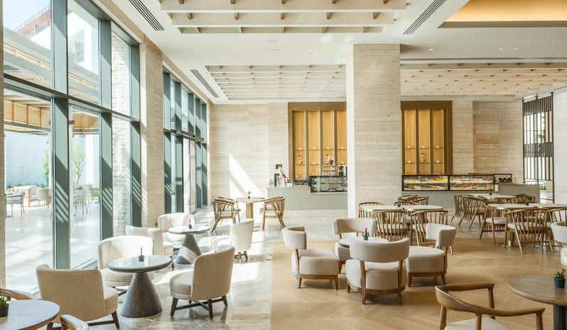 Olivae Patisserie lounge with floor-to-ceiling windows, light wood herringbone floors, and various contemporary seating arrangements under a decorative beamed ceiling at Anda Barut Collection by Kenwood Travel