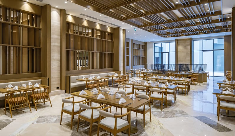 Zestful Bazaar Main Restaurant featuring light wood furniture, marble tile flooring, and a unique geometric wood slat ceiling with warm recessed lighting at Anda Barut Collection by Kenwood Travel