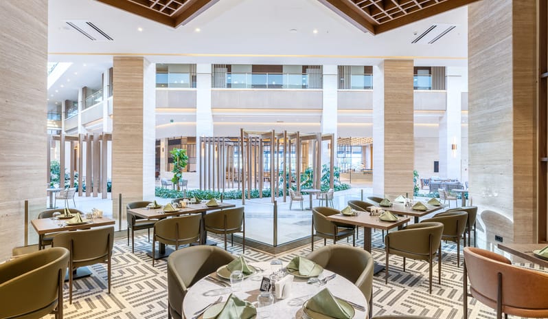 Cibes Restaurant International dining area with marble-topped tables and olive green chairs, featuring an architectural wood lattice ceiling and a view into a multi-story sunlit atrium at Anda Barut Collection by Kenwood Travel