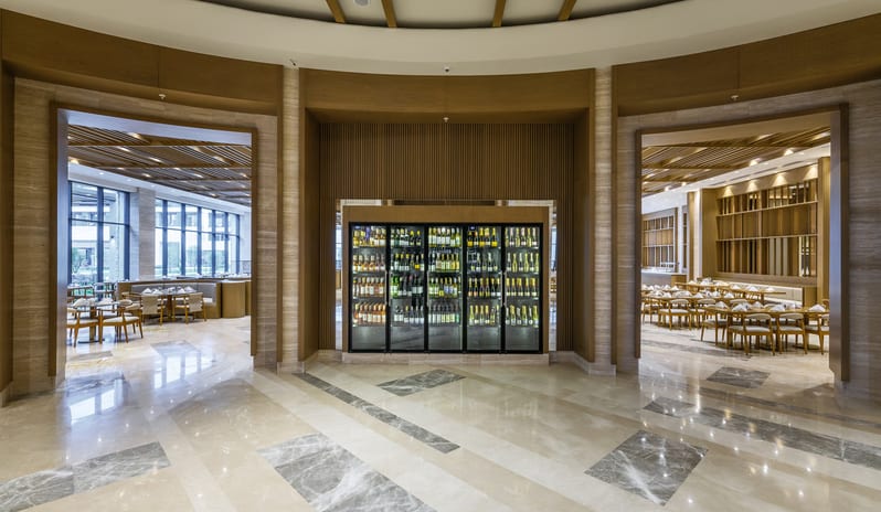 Zestful Bazaar Main Restaurant entrance featuring a large glass wine refrigerator flanked by two open doorways leading to elegant dining areas with marble flooring at Anda Barut Collection by Kenwood Travel