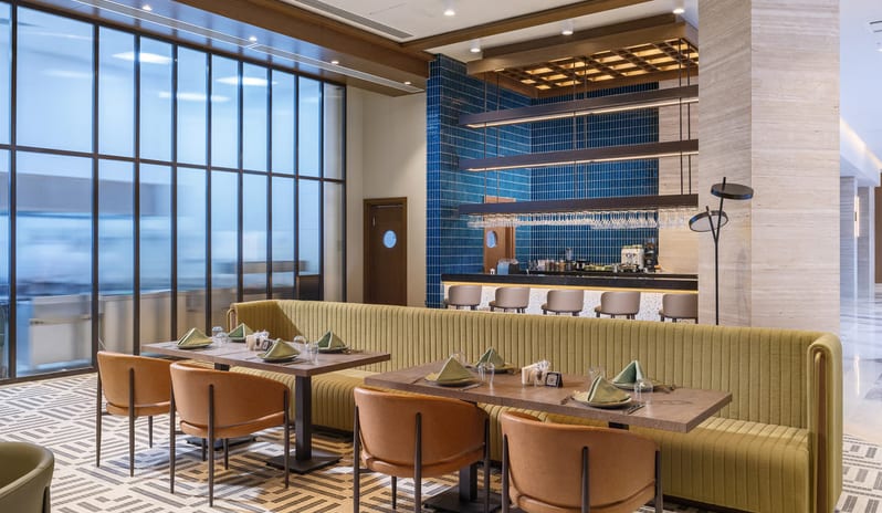 Cibes Restaurant International interior showing a long lime green ribbed banquette booth with wooden tables and tan leather chairs, set against a backdrop of a blue-tiled bar and large frosted glass partitions at Anda Barut Collection by Kenwood Travel